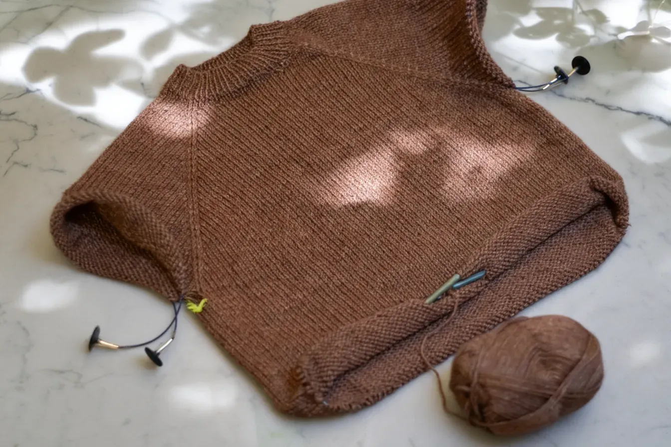 Brown hand-knitted sweater in progress on white marble in dappled sunlight