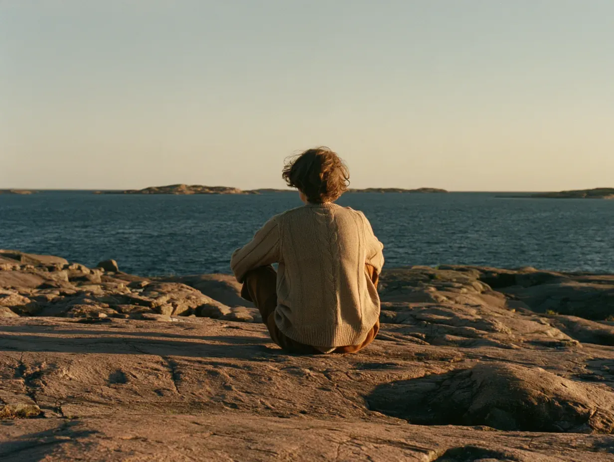 Person in a cream cable-knit sweater sitting on granite rocks facing the sea at golden hour