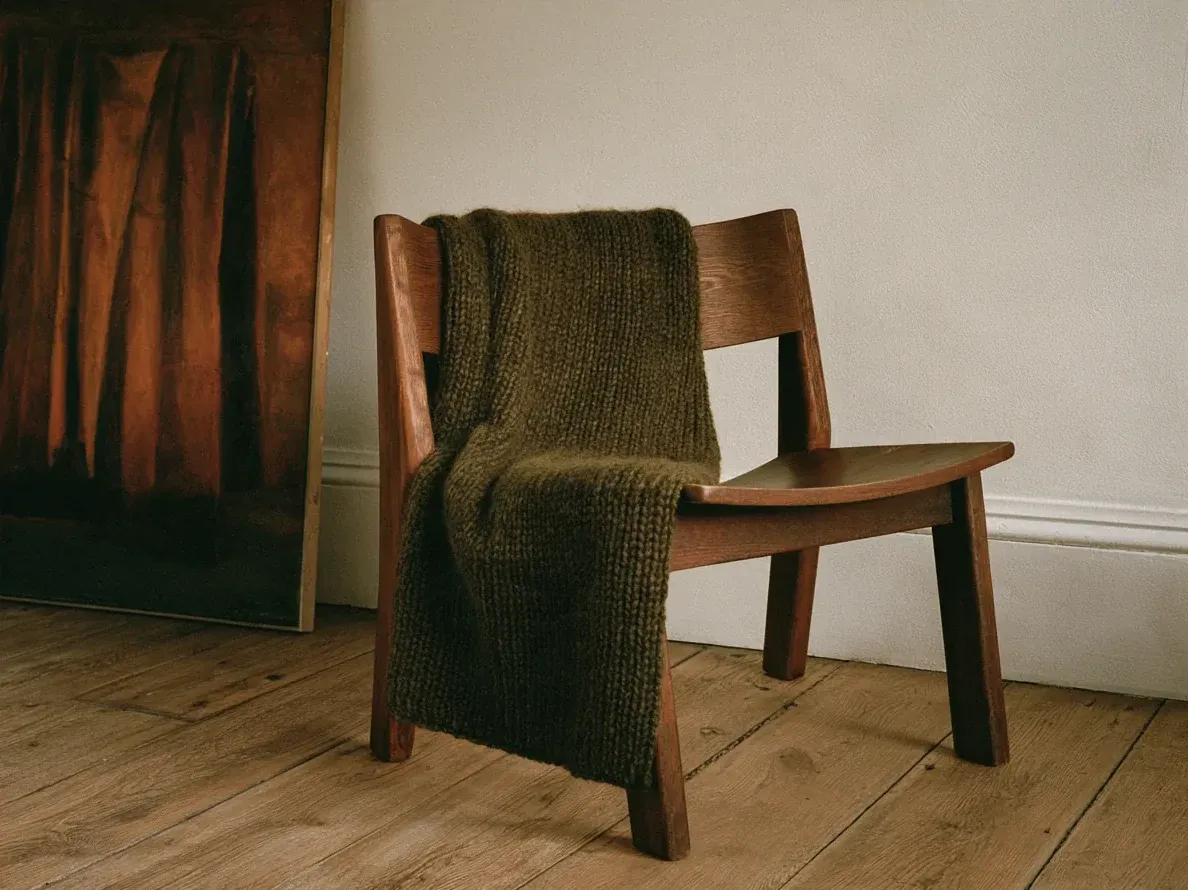 Olive green knitted throw draped over a wooden chair beside a dark painting